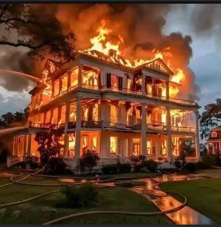 Firefighters spray water on a large white house engulfed in flames and thick smoke.