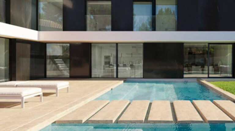 Modern house with glass windows, a pool with stone steps, and white lounge chairs on a beige patio.