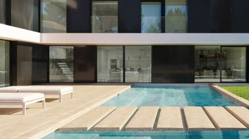 Modern house with glass windows, a pool with stone steps, and white lounge chairs on a beige patio.