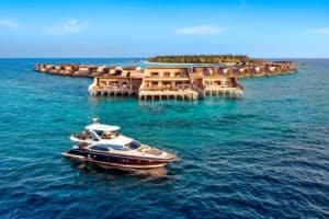 A luxury yacht floats by semi-circular overwater villas on turquoise sea, with a tropical island and blue sky behind.