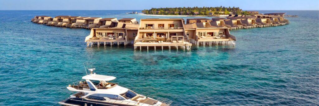 Overwater villas in a circle on clear blue sea, yacht in front, and a tree-covered island in the background.