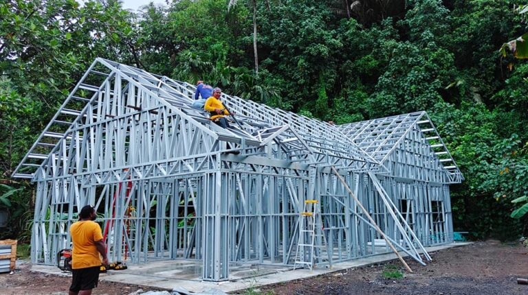 Metal house frame construction in progress with builders working on a lush green outdoor site.