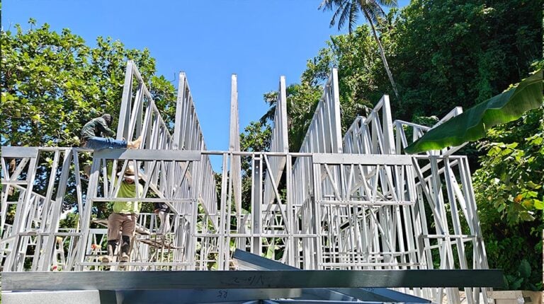 Metal wall frame construction in tropical outdoor setting with lush green foliage and blue sky.