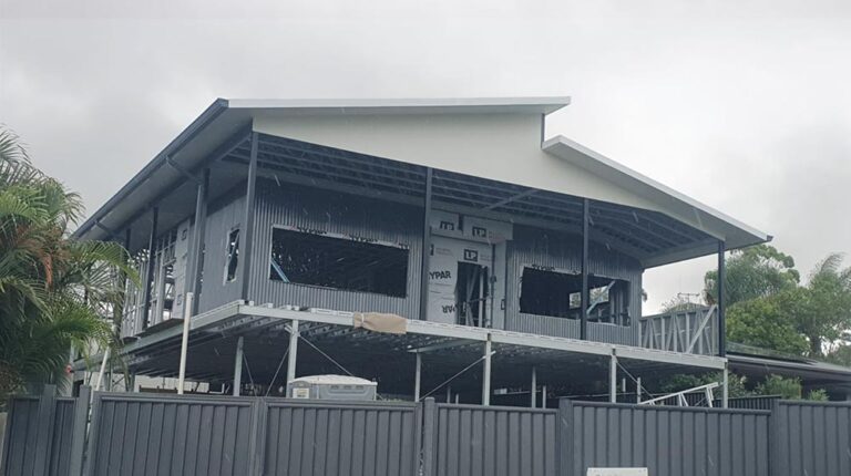 Metal house under construction with a sloped roof, elevated on stilts, surrounded by a metal fence, with construction materials visible, indicating ongoing building process.