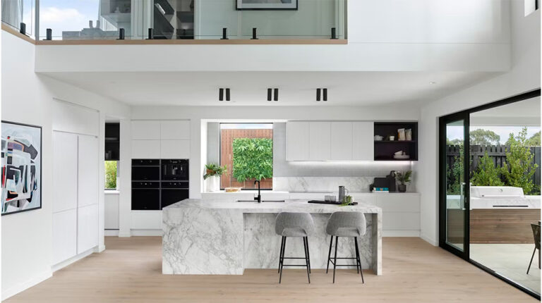 Modern minimalist kitchen with white cabinets, marble island, and large glass sliding door opening to an outdoor patio with hot tub.
