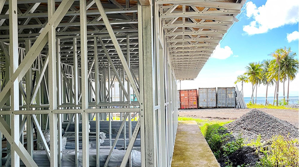 Brightly lit construction site of durable steel framing for a modern building near tropical palm trees and a beach, showcasing high-quality building materials and a sunny maritime environment.