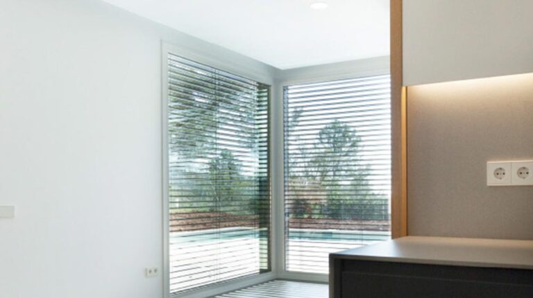 Bright modern corner window with white horizontal blinds and scenic outdoor view of trees and sky, interior home design.