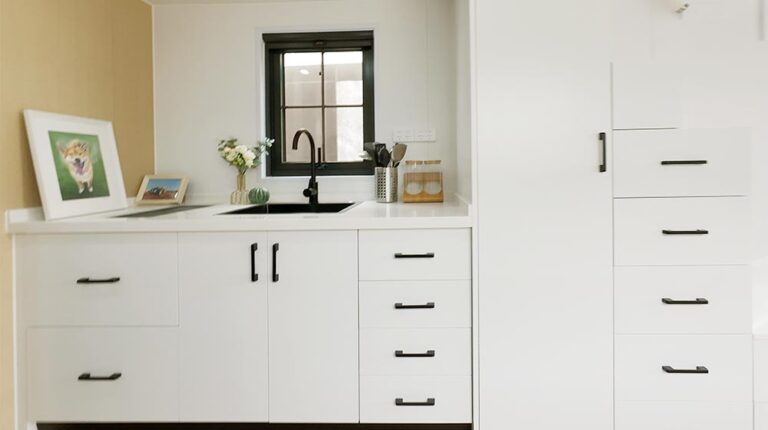 Bright white kitchen cabinets with black handles, a black sink, and a small window, featuring decorative items like a framed painting, flowers, and kitchen tools for a clean, fresh look.