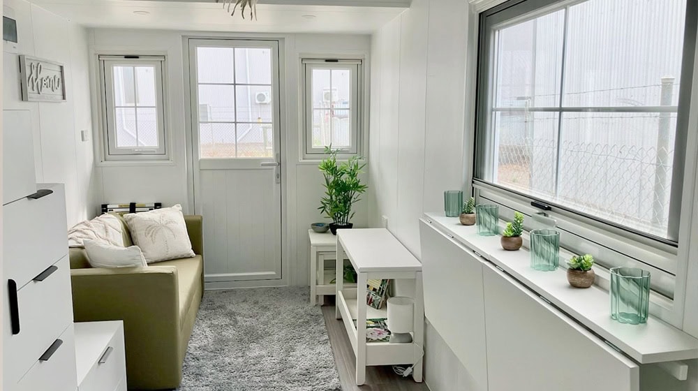 Bright modern sunroom with white walls and large windows, featuring a beige sofa, green plants, and decorative glass vases, perfect for relaxing or indoor gardening.