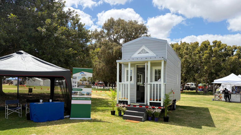 A stylish, compact tiny house on wheels set on a grassy field, surrounded by trees and vendor tents, showcasing innovative sustainable living solutions from DeepBlueHome.