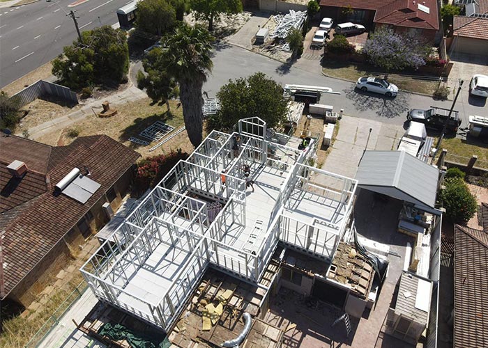Aerial view of a residential home under construction, showcasing a steel framework structure with surrounding trees, nearby houses, and construction materials, highlighting innovative building techniques.
