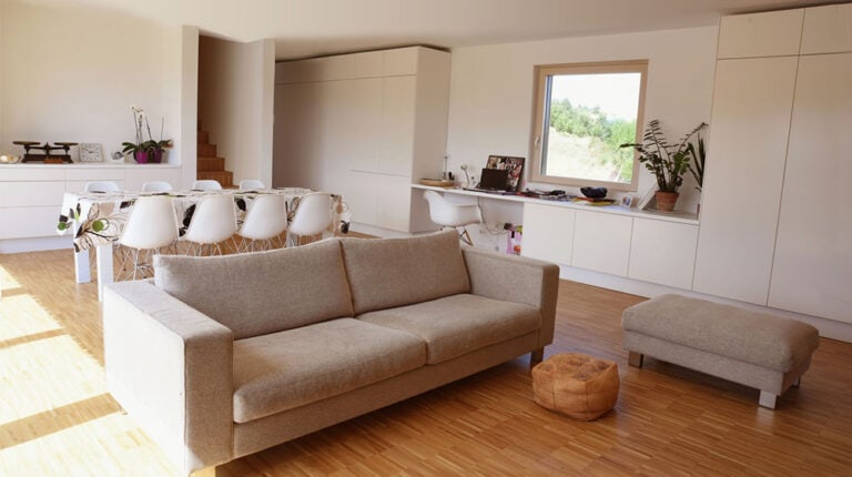 Bright, airy living room featuring a beige sofa, white cabinetry, and large window with greenery view.