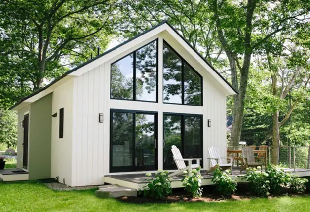A-Frame tiny house with expansive glass windows, white exterior, and outdoor deck, nestled in a lush green forest setting. Perfect for sustainable living and nature retreats.