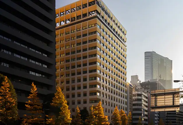 Office building with yellow and gray exterior in downtown city skyline, surrounded by other high-rise structures and trees, under a clear sky.
