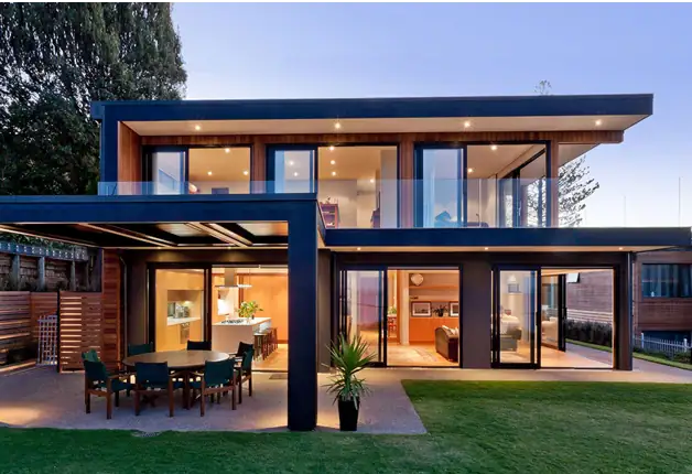 Modern two-story house featuring expansive glass windows, a spacious outdoor patio with dining area, and contemporary architectural design emphasizing natural light and outdoor living.