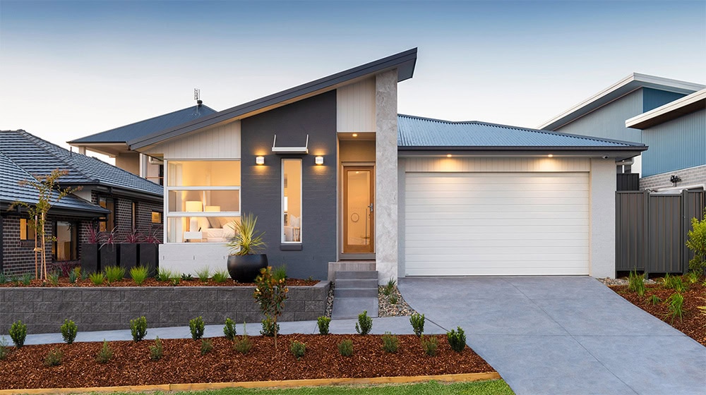 A contemporary single-story home featuring a stylish sloped roof, large windows, and a spacious garage. The front yard is landscaped with small shrubs, mulch, and a paved driveway, creating an invitin.