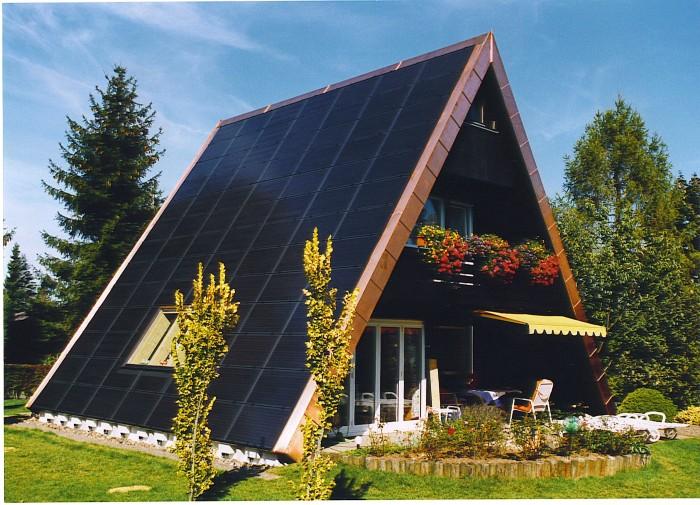 Modern A-frame house with extensive solar panel roofing, surrounded by greenery, featuring a balcony with flowers and outdoor seating, emphasizing sustainable living and eco-friendly design.