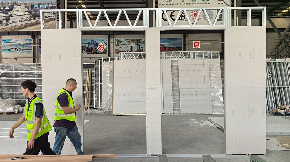 Two construction workers in safety vests walking through a building site with white wall panels and construction materials in the background.