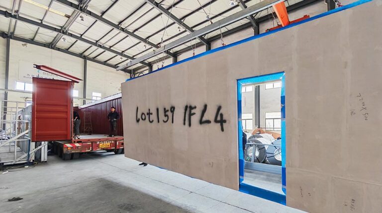 Construction site showing a large panel with blue window frame and graffiti, inside a warehouse with a truck and workers.