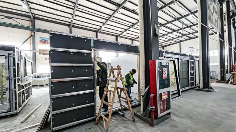 Workers assembling a modular prefab house in a spacious industrial warehouse with steel framework and construction materials.