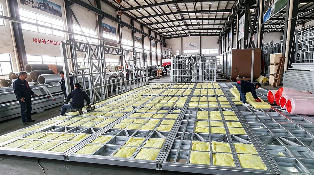 Workers assembling the metal framework and insulation panels for a prefab house in a factory setting.