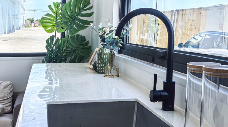 Contemporary kitchen sink with sleek black faucet, white marble countertop, and decorative plants near a large window, showcasing modern home design.