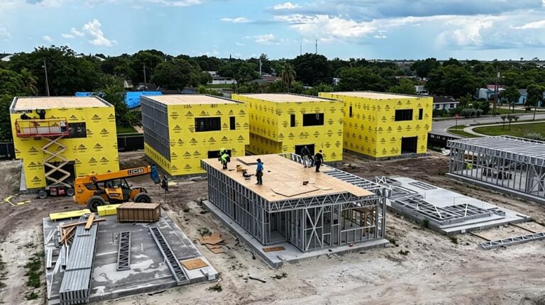 Construction site with yellow and gray building frames, construction workers, and equipment under a partly cloudy sky.