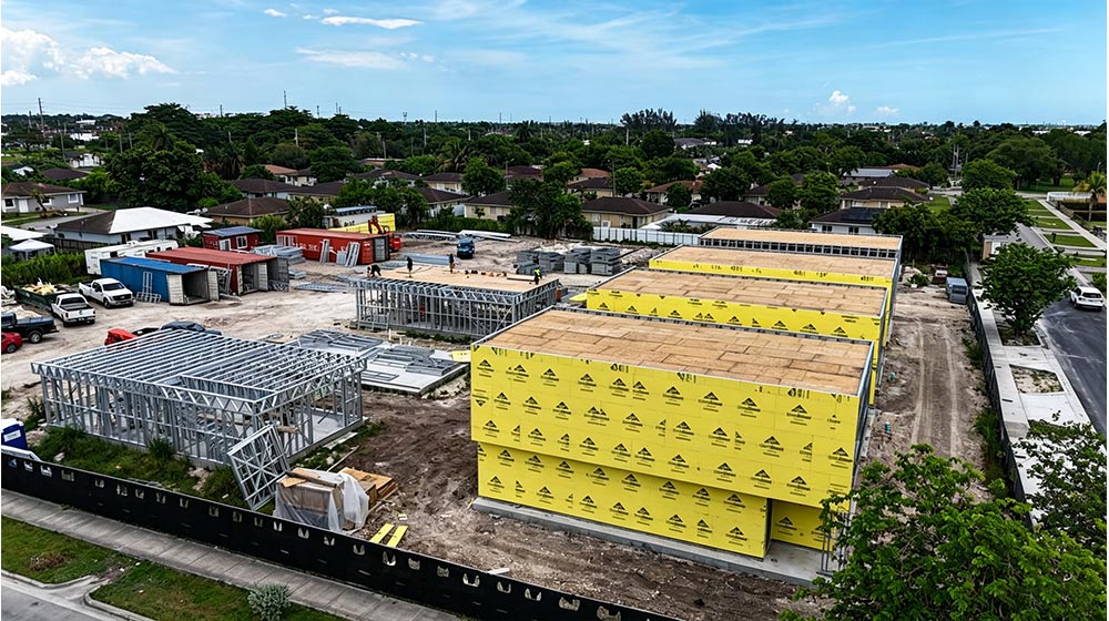 Aerial view of a construction site with yellow insulation panels, steel framework, and surrounding residential area under a partly cloudy sky.