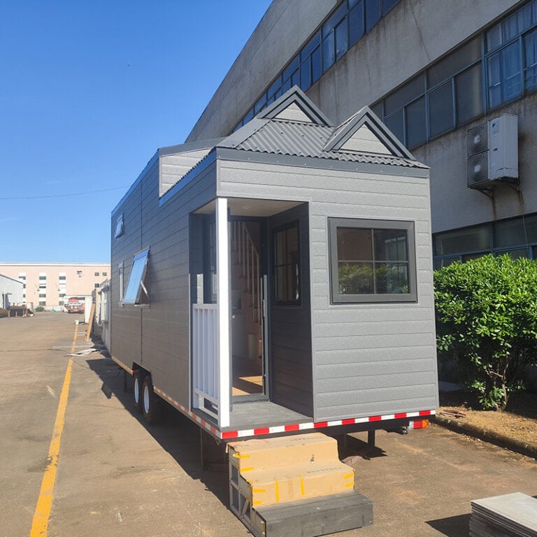 Modern tiny house on wheels with gray exterior, small steps, and a front porch, parked outdoors near a large building.