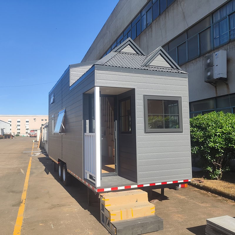 Modern tiny house on wheels with gray exterior, small steps, and a front porch, parked outdoors near a large building.