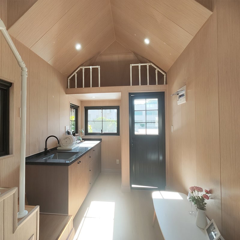 Interior view of a compact, modern tiny house kitchen with wooden walls, black countertop, and a small window providing natural light.