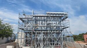 Construction site with metal scaffolding and framework for a building, under a partly cloudy sky, indicating ongoing development.