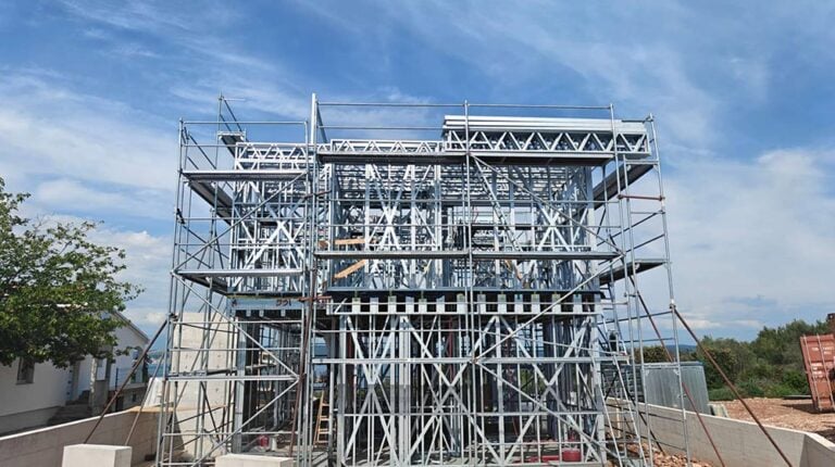 Construction site with metal scaffolding and framework for a building, under a partly cloudy sky, indicating ongoing development.