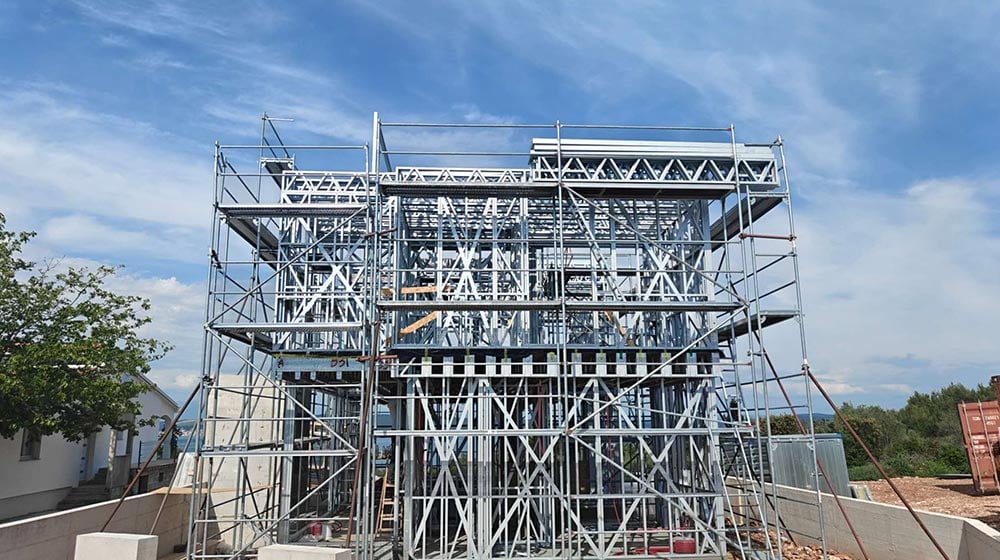 Construction site with metal scaffolding and framework for a building, under a partly cloudy sky, indicating ongoing development.