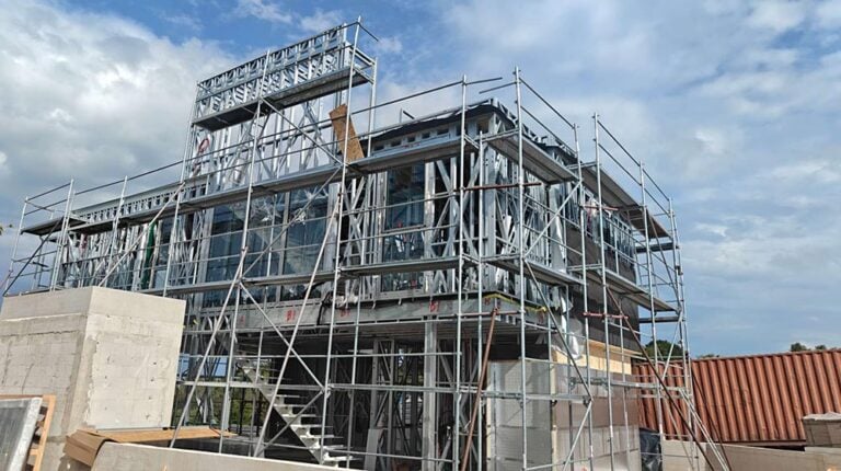 Construction site showing a multi-story building with metal scaffolding and partially installed glass windows under a partly cloudy sky.