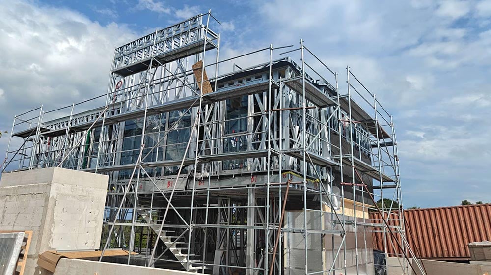 Construction site showing a multi-story building with metal scaffolding and partially installed glass windows under a partly cloudy sky.