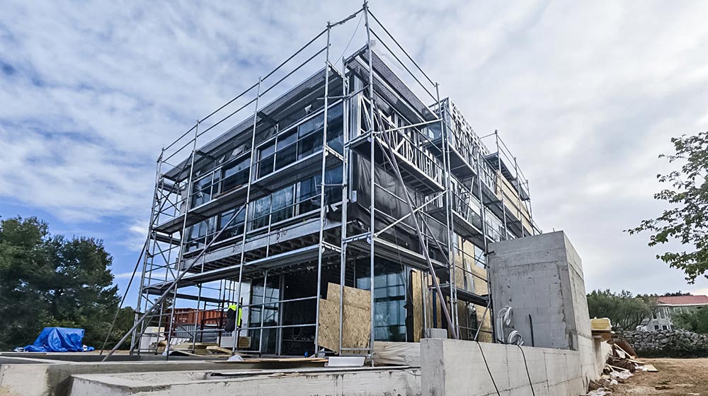 Construction site featuring a multi-story building with scaffolding, safety barriers, and construction materials, set against a partly cloudy sky.