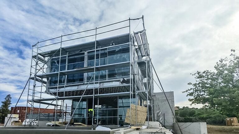 Construction site featuring a modern glass building with scaffolding and safety barriers, surrounded by trees and a partly cloudy sky.