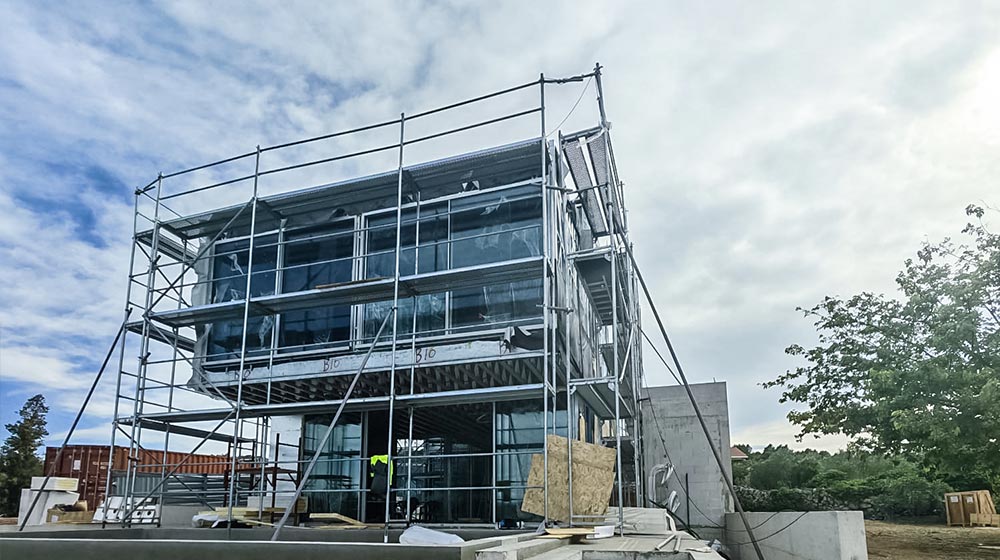 Construction site featuring a modern glass building with scaffolding and safety barriers, surrounded by trees and a partly cloudy sky.