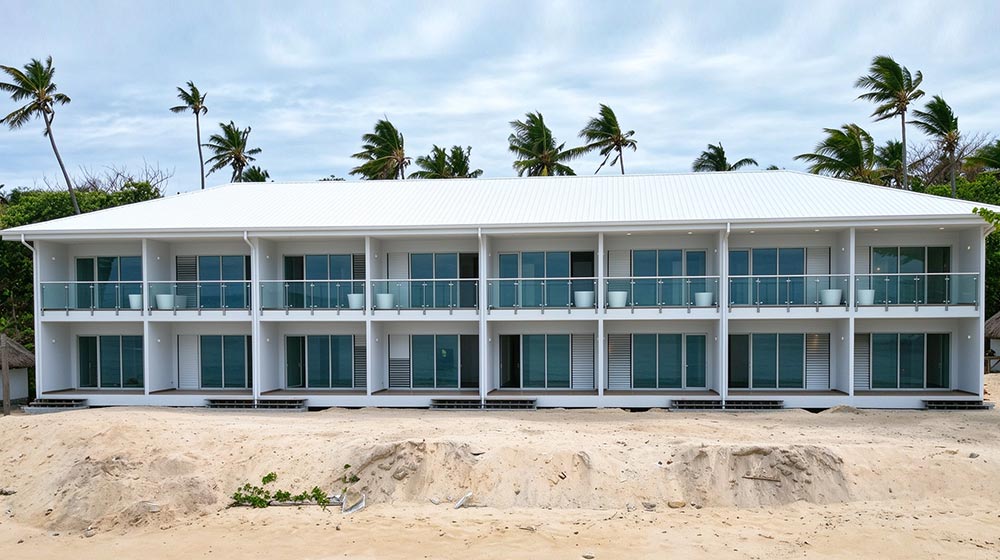 Contemporary beachfront hotel with multiple rooms and balconies, surrounded by palm trees and sandy beach, under a partly cloudy sky.