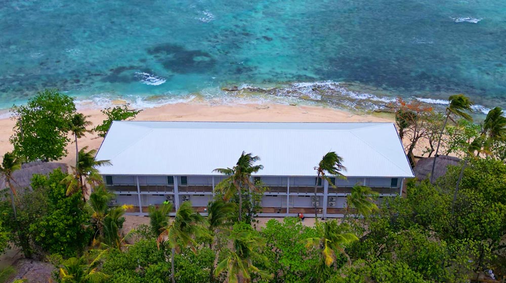 A beachfront hotel in Fiji featuring a light steel frame structure, surrounded by lush greenery and overlooking the clear blue ocean, ideal for vacation stays.
